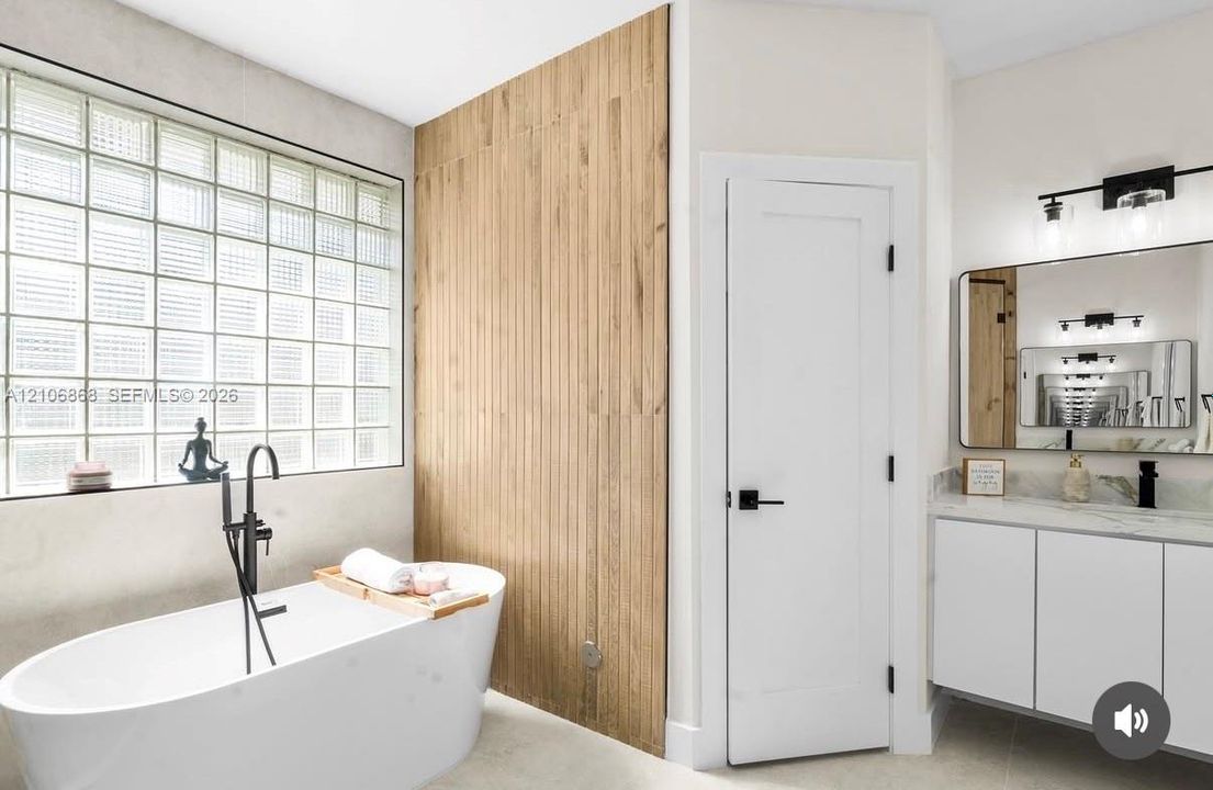 Freestanding soaking tub with matte black floor-mounted faucet, glass block window, wood-finish vertical accent wall, floating vanity with stone countertop, and black hardware finishes