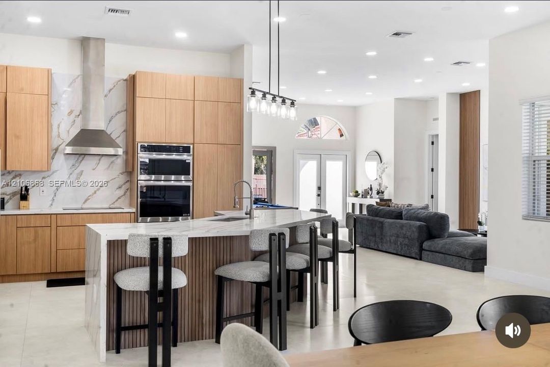 Kitchen featuring flat-panel wood cabinetry, stone-patterned backsplash, built-in double wall ovens, stainless steel vent hood, and an expansive waterfall-edge island with vertical wood detailing