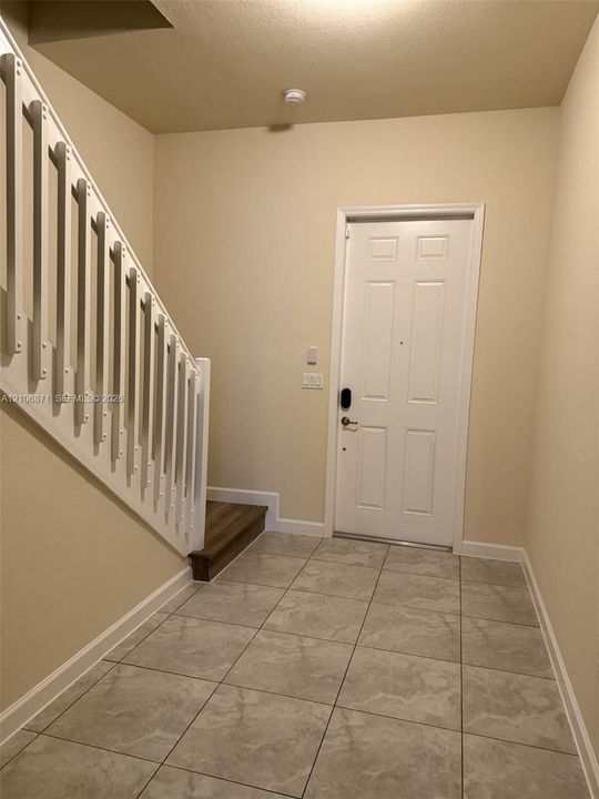 Entryway featuring large-format ceramic tile flooring, a six-panel exterior door with electronic deadbolt, and a staircase with white balusters and wood-finish tread