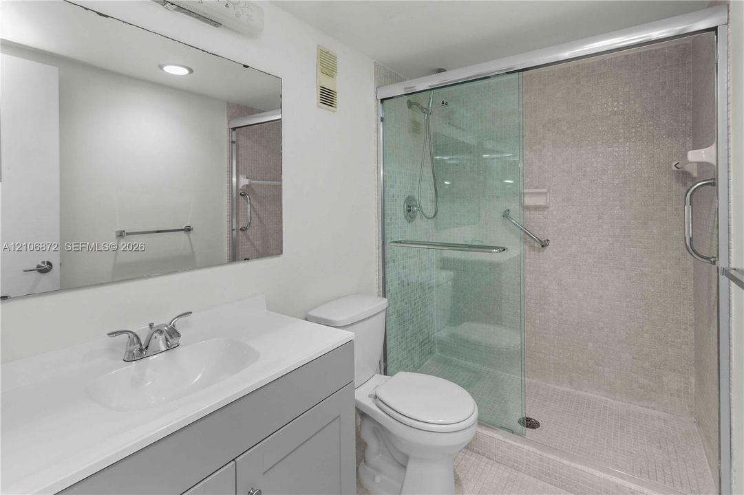 Bathroom featuring a vanity with a solid-surface countertop, an oversized wall mirror, a walk-in shower with glass sliding doors, and mosaic tile shower wall cladding