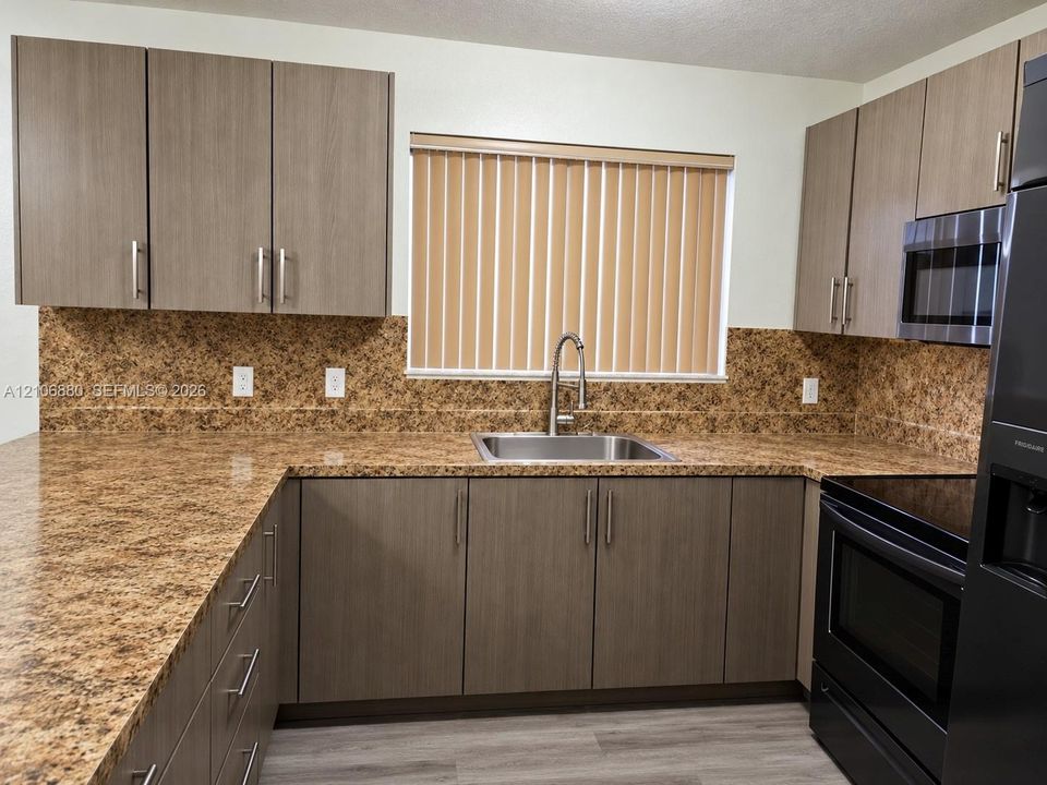 Kitchen featuring flat-panel wood-finish cabinetry, stone-patterned countertops with matching backsplash, stainless steel sink with pull-down faucet, and built-in black appliances