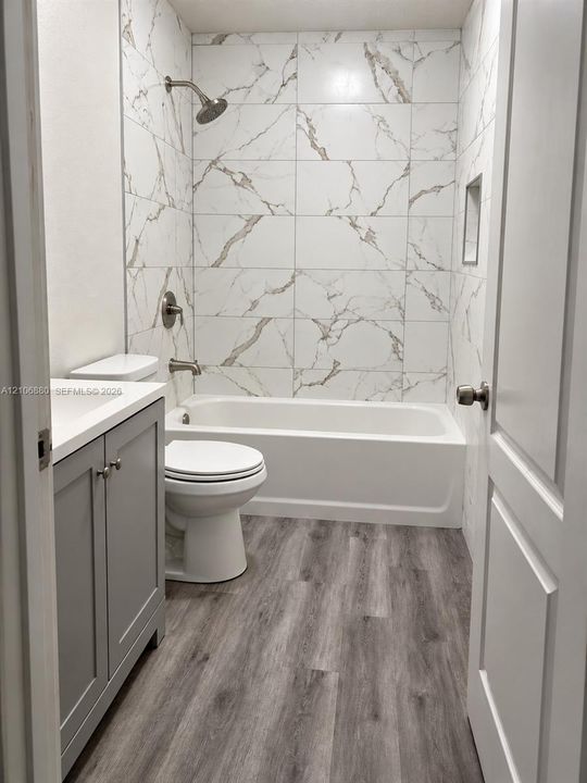 Bathroom featuring a vanity with a light-toned countertop, wood-finish flooring, and a tub-shower combination enclosed by large-format tile with decorative veining and a recessed shower niche