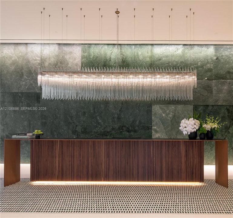 Large-scale linear chandelier with crystalline fringe, wood-veneer floating desk with base accent lighting, and full-height green-toned stone feature wall