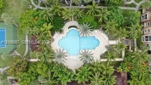 Aerial perspective of a common area featuring a swimming pool, paved deck, hard-surface tennis court, and lush landscaping with mature palm trees