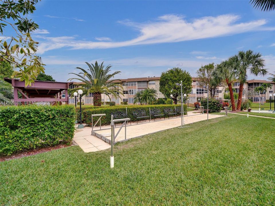 Outdoor recreational space featuring a bocce ball court, manicured hedges, mature palm trees, and structural shaded pavilion