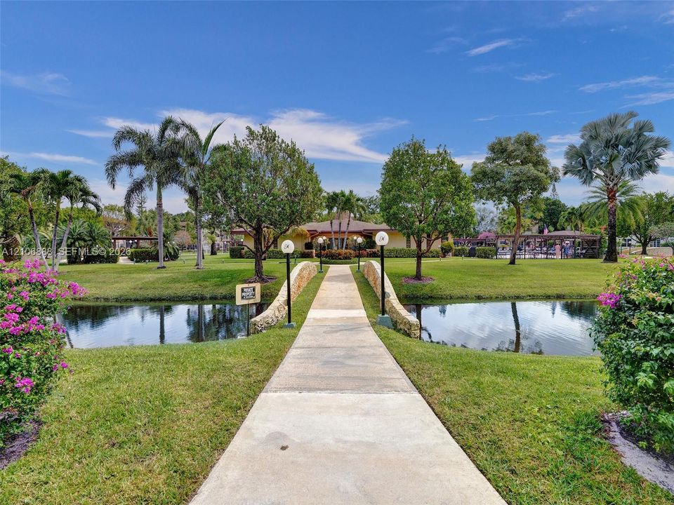 Concrete walkway featuring a stone-clad bridge crossing a water feature, surrounded by manicured landscaping and mature trees