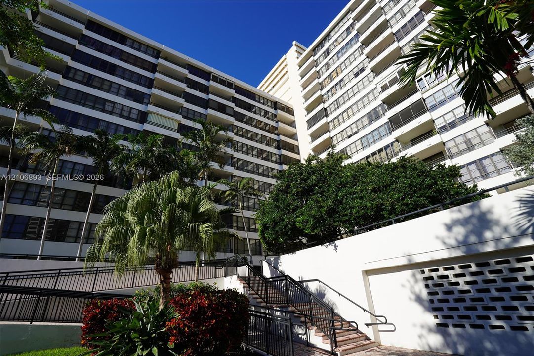 Multi-story residential building featuring private balconies, concrete construction, decorative masonry screening, and exterior stairways framed by mature palms and landscaping