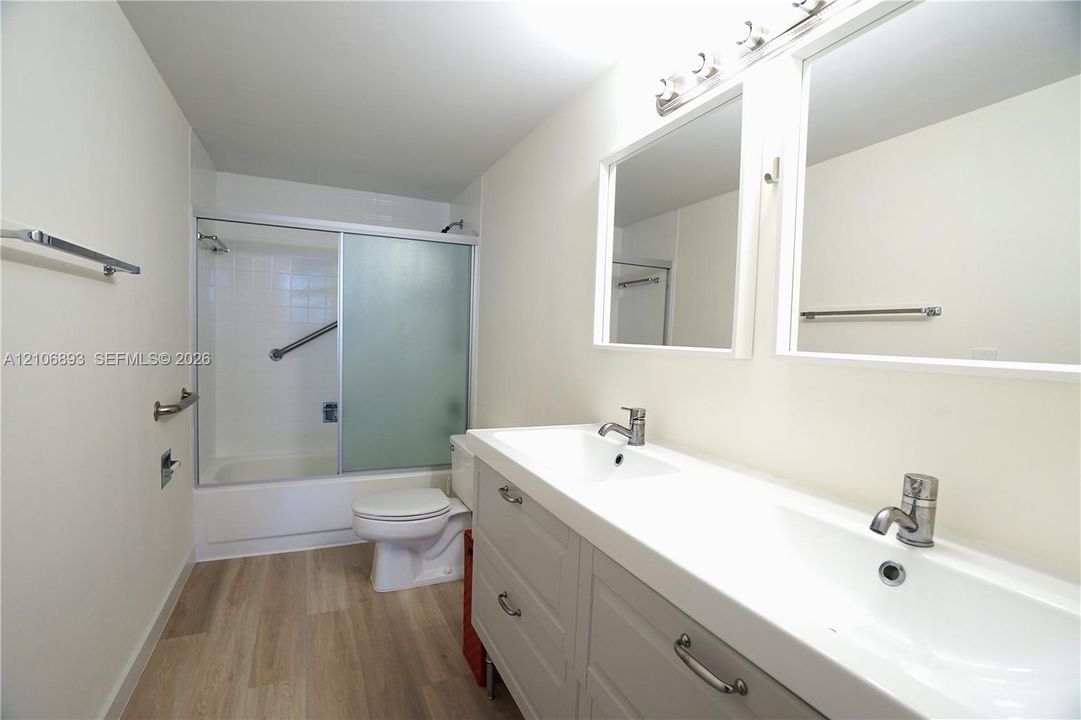 Bathroom featuring a dual-sink vanity with brushed metal hardware, wood-finish flooring, two large wall-mounted mirrors, and a tub-shower combination with sliding glass doors