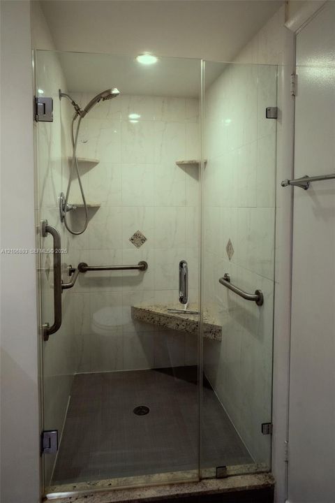 Walk-in shower featuring a frameless glass enclosure, stone-surface corner bench, adjustable handheld showerhead, wall-mounted support rails, and tiled surround