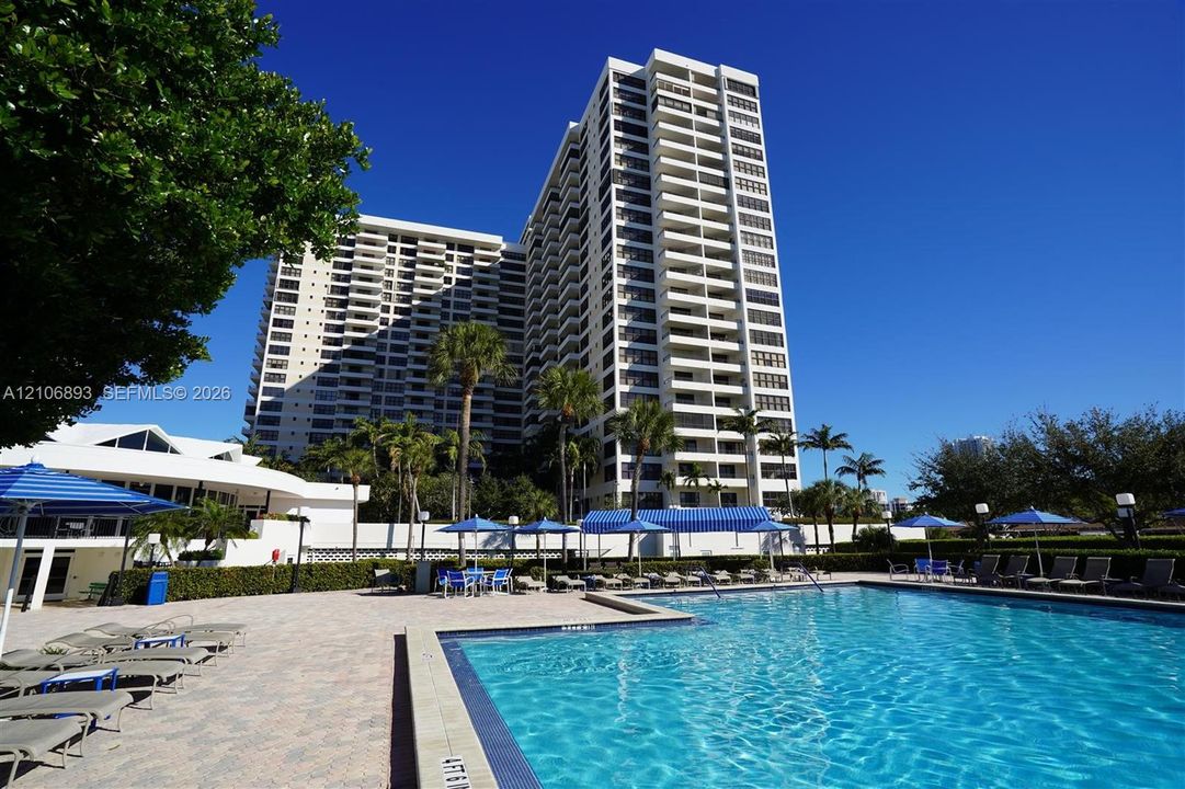 Multi-story high-rise building featuring private balconies, expansive swimming pool with tiled edge, stone paver pool deck, and landscaped grounds with mature palm trees
