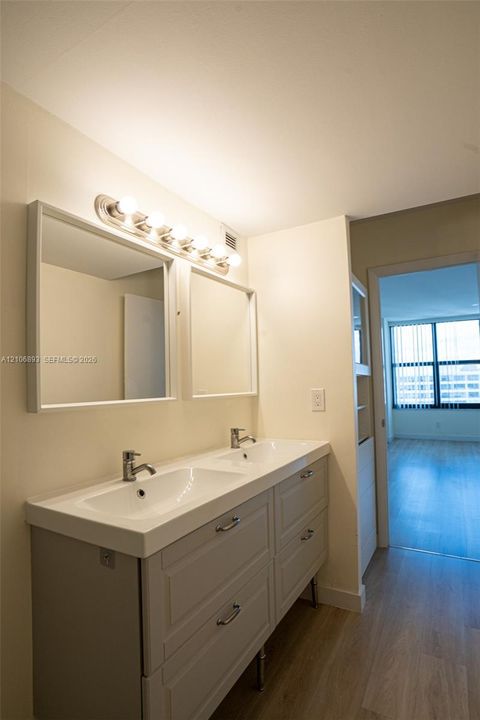 Double vanity featuring integrated sinks, dual rectangular wall-mounted mirrors, brushed-nickel hardware, and a multi-bulb light fixture, paired with wood-finish flooring