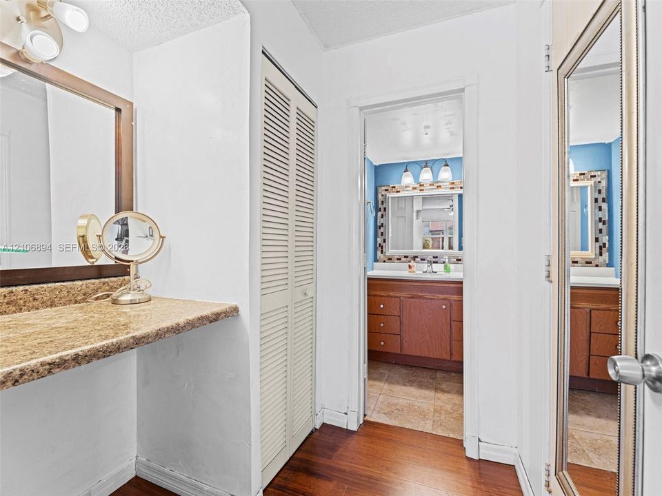 Dressing area featuring stone-finish countertop, wood-finish flooring, and floor-to-ceiling louvered closet doors, leading to a vanity area equipped with wood cabinetry and tile flooring