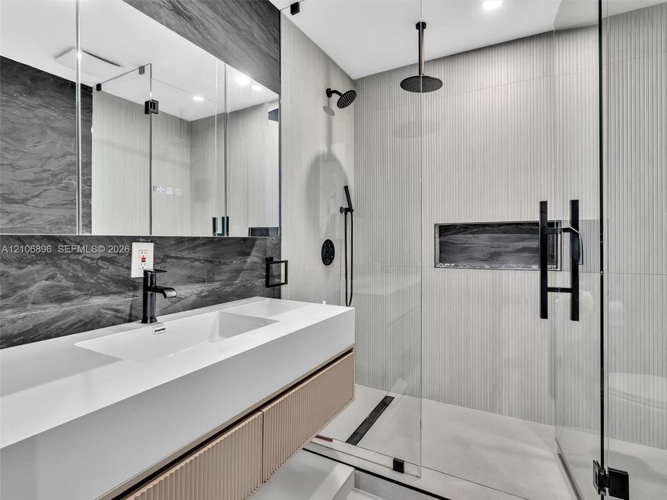 Floating vanity with an integrated sink and textured wood-finish cabinetry, paired with a frameless glass shower enclosure featuring matte black rainfall and handheld showerheads, a recessed wall niche, and large-format wall tile
