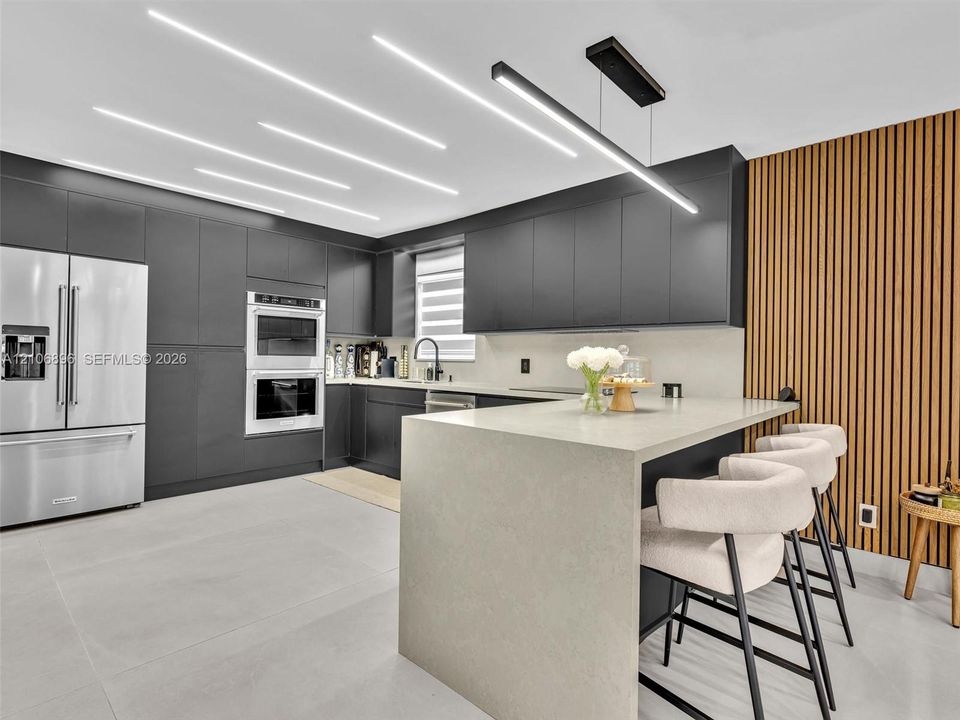 Modern kitchen featuring flat-panel cabinetry, a waterfall-edge stone island, linear LED ceiling lighting, integrated double wall ovens, and a vertical wood-slat accent wall