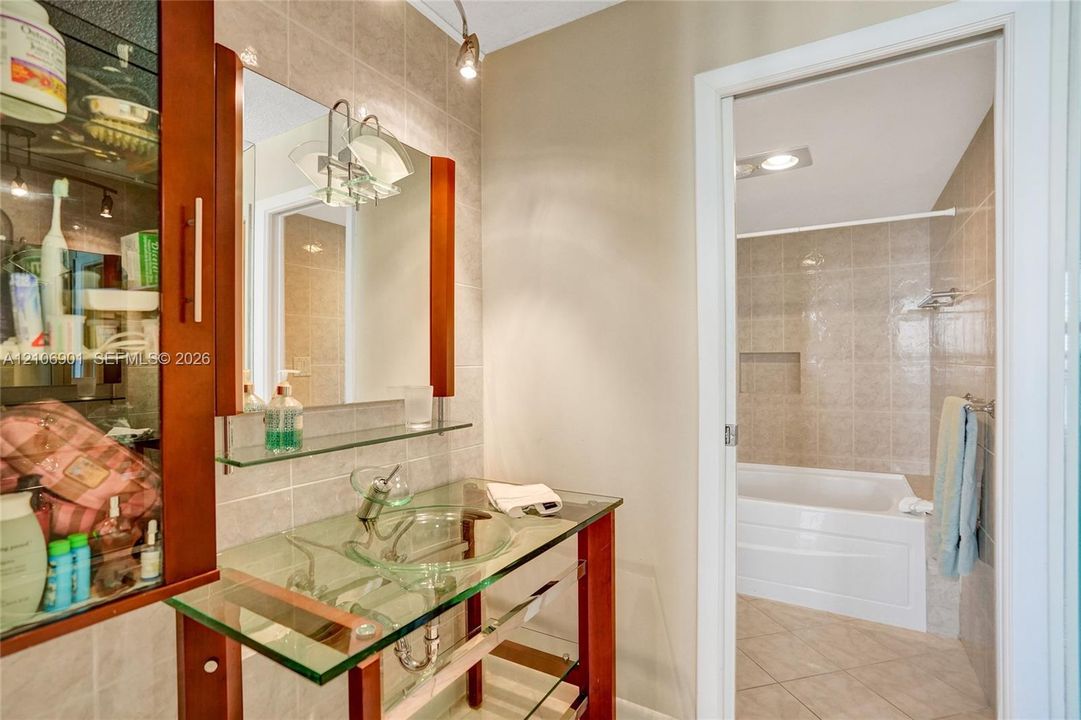 Vanity featuring a glass countertop, integrated vessel sink, and wood-finish frame, complemented by a wall-mounted mirror, glass shelving, and tile shower surround with a built-in recessed niche