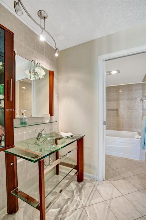 Glass vessel sink vanity featuring a wood-finish frame, clear glass shelving, wall-mounted track lighting, and a ceramic-tiled tub enclosure
