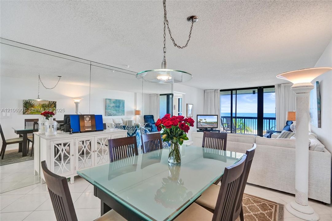 Open-concept main living area featuring large-format ceramic tile flooring, a wall-to-wall mirrored accent wall, suspended glass-shade pendant lighting, and a floor-to-ceiling glass slider with balcony access