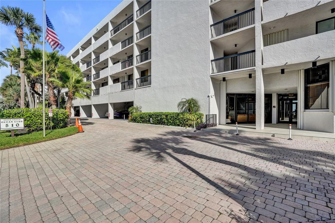 Multi-story concrete exterior with private balconies and metal railings, paver-stone driveway, and manicured tropical landscaping
