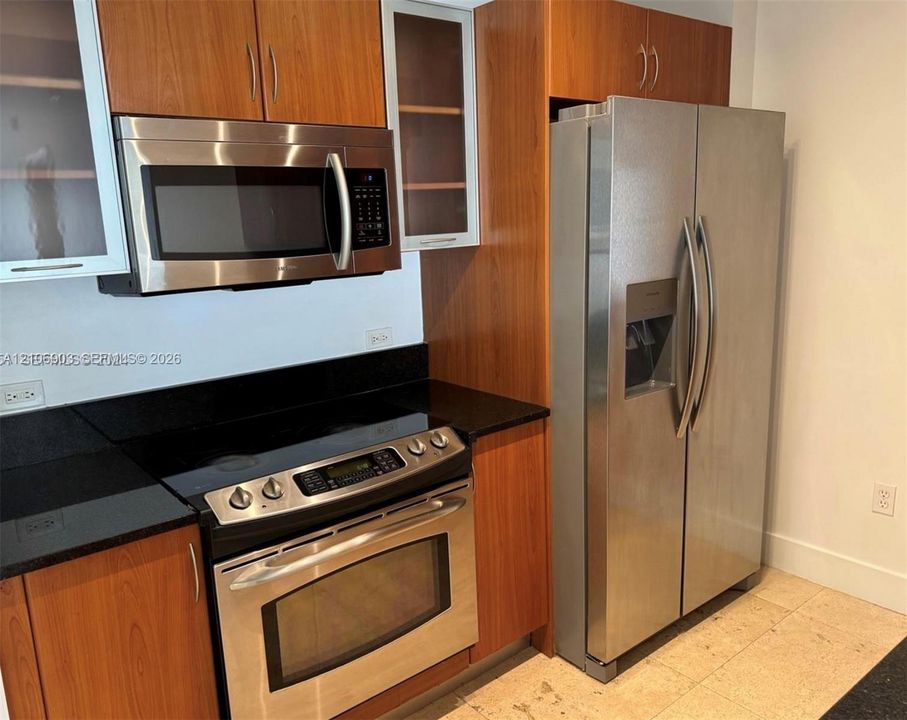 Kitchen featuring wood-veneer cabinetry with glass-panel upper cabinet doors, stone-finish countertops, and stainless steel appliances including a side-by-side refrigerator, over-the-range microwave, and slide-in electric range