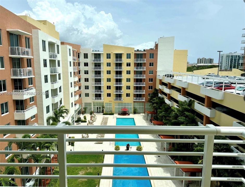 Multi-story residential complex featuring private balconies, a rectangular swimming pool, landscaped green space, and a structured parking facility