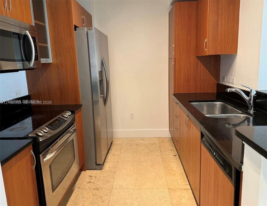 Galley kitchen featuring wood-finish cabinetry, dark stone countertops, stainless steel appliances, a dual-basin undermount sink, and tile flooring