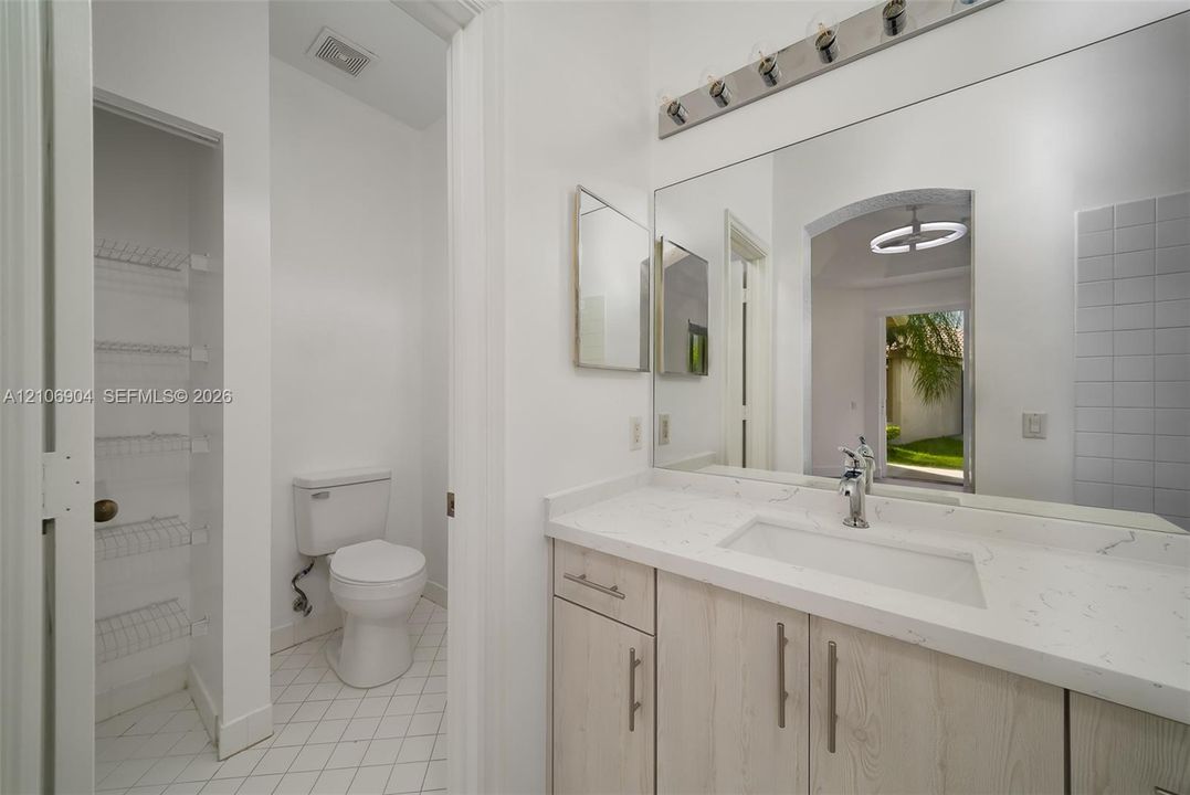 Vanity featuring a light wood-finish cabinet, quartz countertop, and integrated sink, complemented by a large wall-mounted mirror, multi-bulb lighting fixture, and built-in shelving