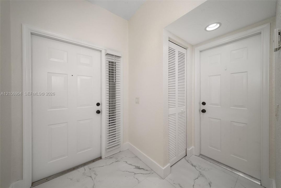 Entryway featuring polished marble-style tile flooring, two six-panel doors with matte black hardware, vertical slatted window blinds, and a built-in louvered closet door with recessed ceiling lighting