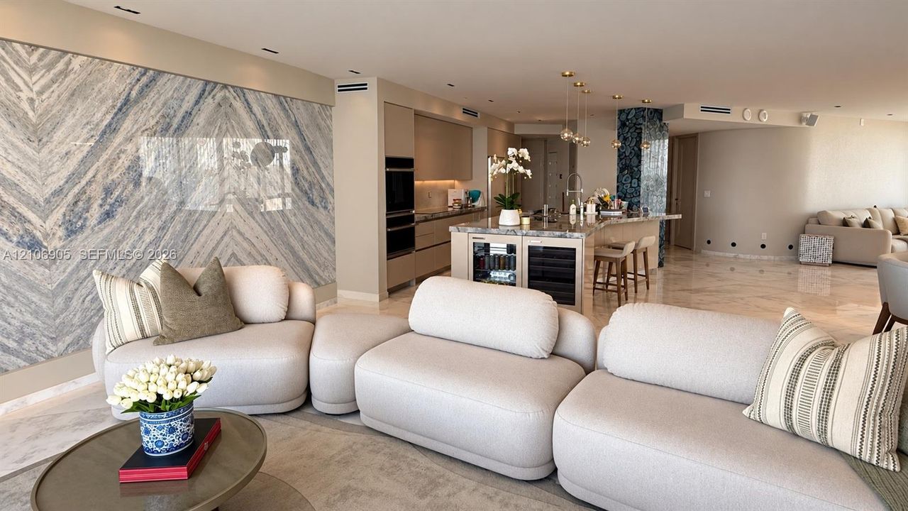 Open-concept living space featuring a floor-to-ceiling stone accent wall, natural stone flooring, an expansive kitchen island with a built-in beverage center, and recessed lighting