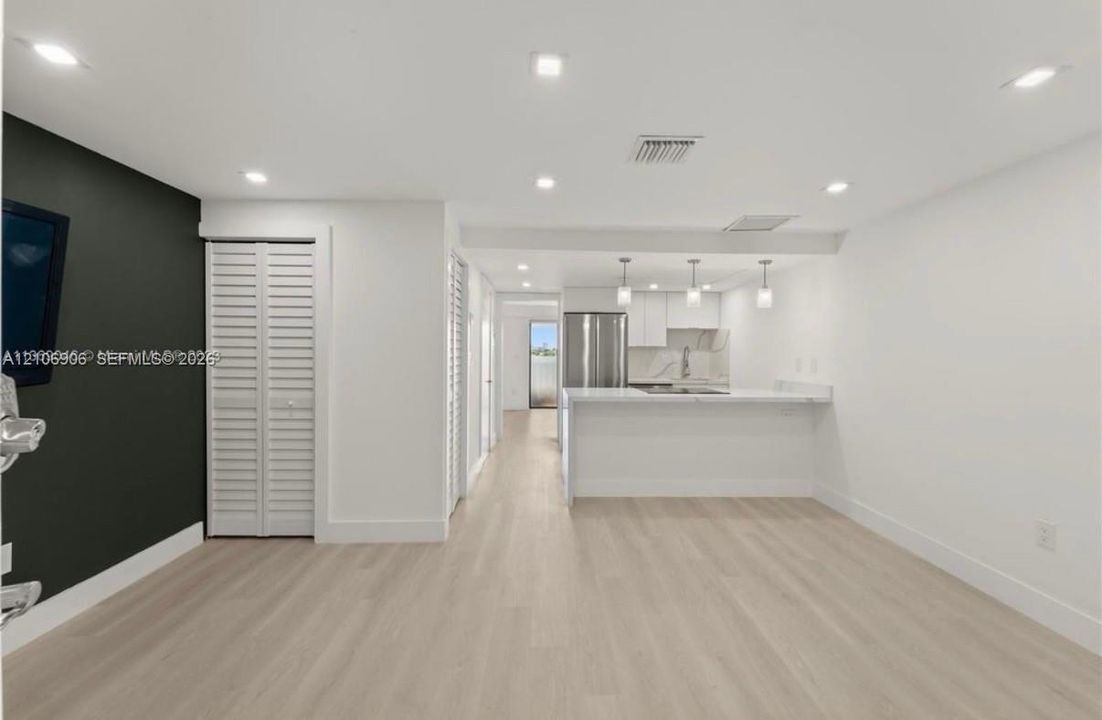 Open-concept space featuring light wood-finish flooring, recessed lighting, white flat-panel cabinetry, stone-surfaced breakfast bar, and stainless steel appliances
