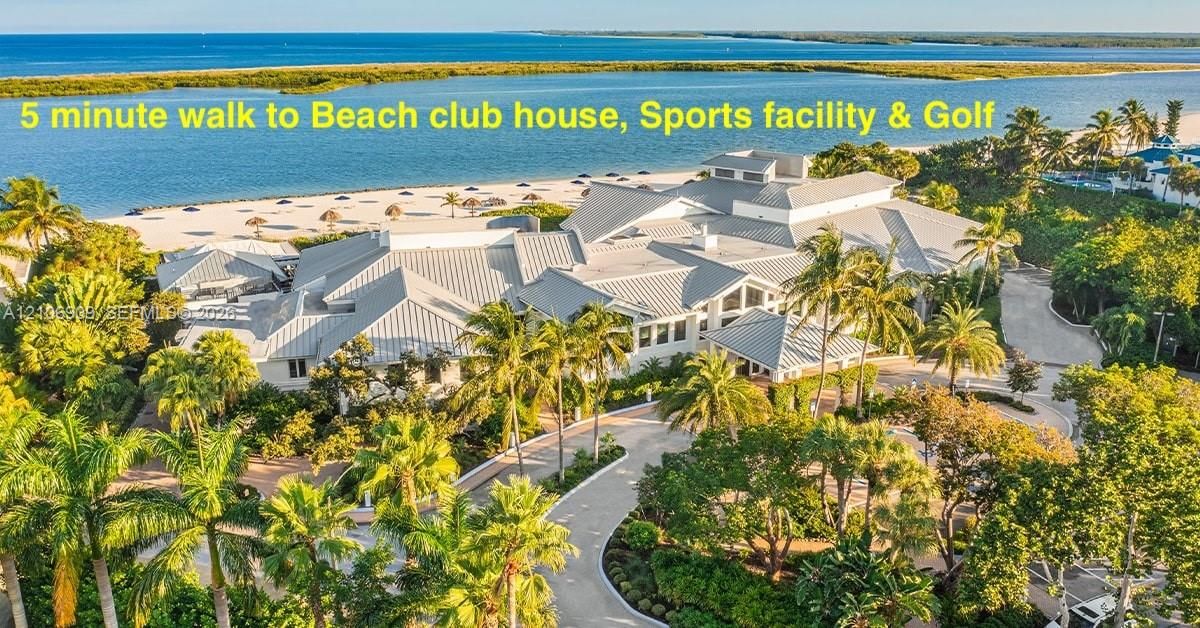 Coastal clubhouse featuring a metal roof, paved circular drive, expansive manicured landscaping, and direct proximity to a sandy shoreline