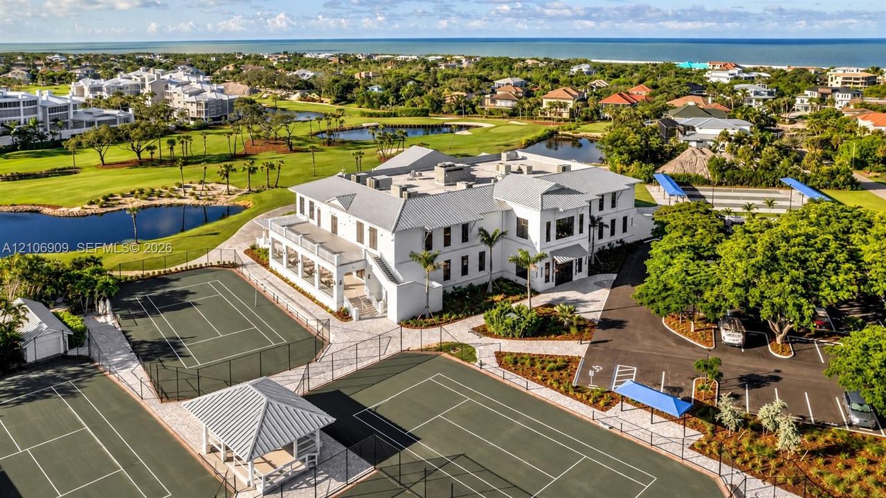 Aerial perspective of a white multi-story exterior featuring multiple tennis courts, a covered pavilion, asphalt parking, and manicured landscaping
