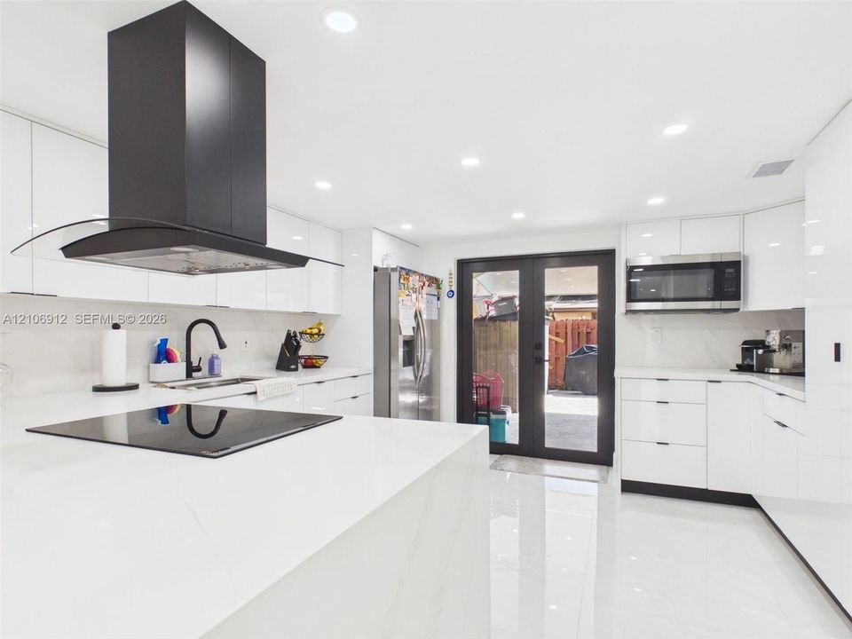 Modern kitchen featuring high-gloss white cabinetry, expansive stone countertops, a stainless steel built-in microwave, a matte black range hood, and glass-paneled double doors