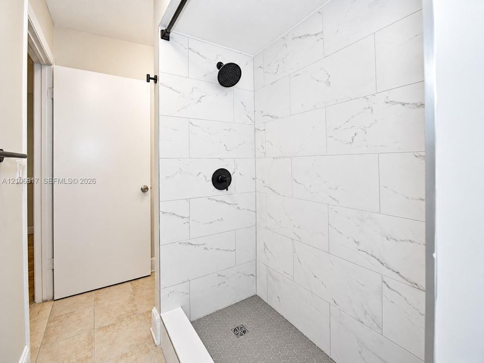 Walk-in shower featuring large-format white stone-finish wall tile, grey hexagonal floor tile, matte black wall-mounted shower head, and matte black single-handle pressure balance valve
