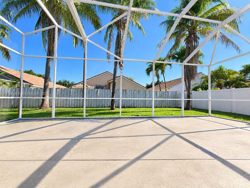 Screen-enclosed concrete patio featuring a white metal frame, privacy fencing, and mature palm trees