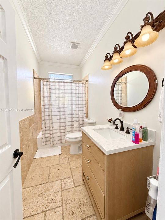Bathroom featuring a vanity with a light-finish wood cabinet and a stone-look countertop, stone tile flooring, wall-mounted three-light fixture, and a tub-shower combination with floor-to-ceiling stone tile surround