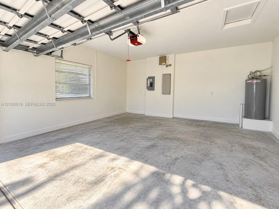 Finished garage interior featuring smooth-finish concrete flooring, painted walls, an automatic overhead door opener, a window with horizontal blinds, and an integrated water heater
