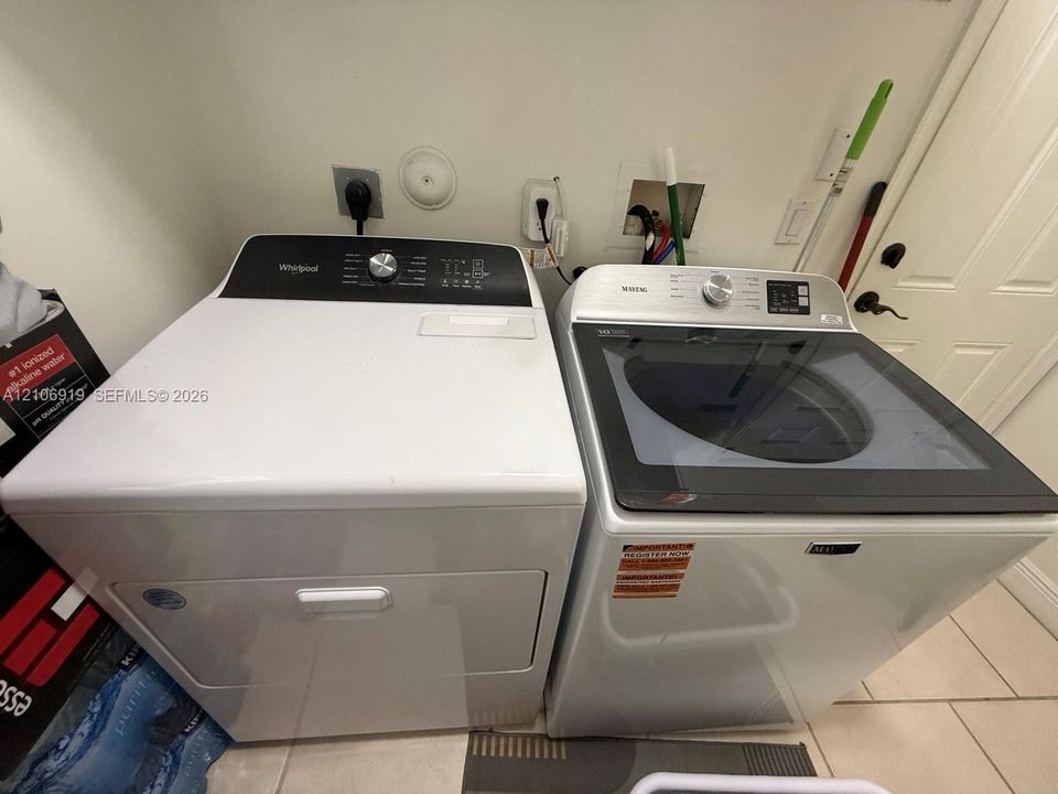 Laundry area featuring a side-by-side Whirlpool dryer and Maytag top-load washer with glass lid, set against a neutral wall with ceramic tile flooring