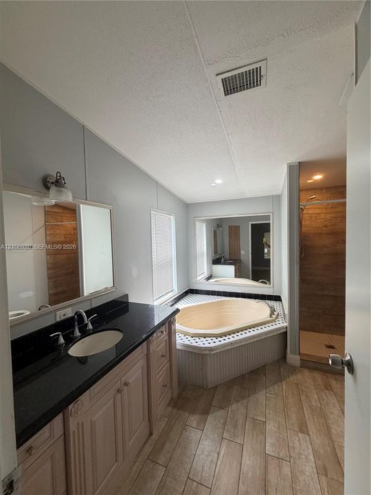 Bathroom featuring wood-finish flooring, a vanity with a dark countertop and integrated sink, a deep soaking tub with a tiled surround, and a walk-in shower with wood-patterned wall tile