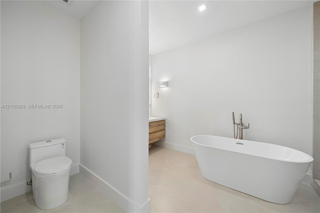 Freestanding white soaking tub with polished chrome floor-mounted faucet, wood-finish vanity with countertop basin, and low-profile elongated toilet