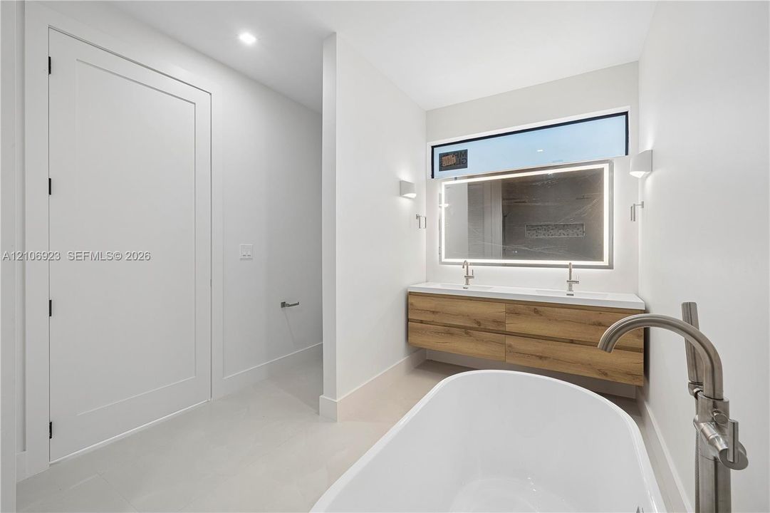 Freestanding soaking tub with floor-mounted brushed nickel faucet, floating wood-finish vanity with dual integrated sinks, and oversized LED-backlit mirror