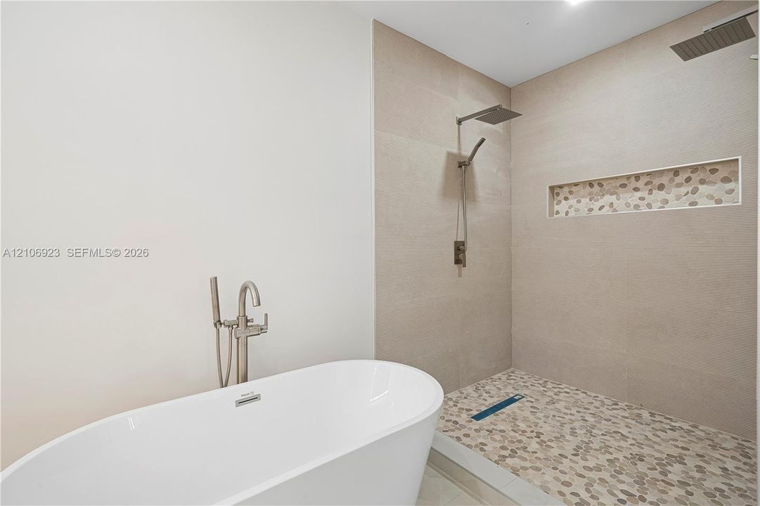 Freestanding soaking tub with a floor-mounted brushed nickel faucet, walk-in shower featuring large-format wall tile, pebble-stone flooring, rainfall showerhead, and a built-in tiled shower niche