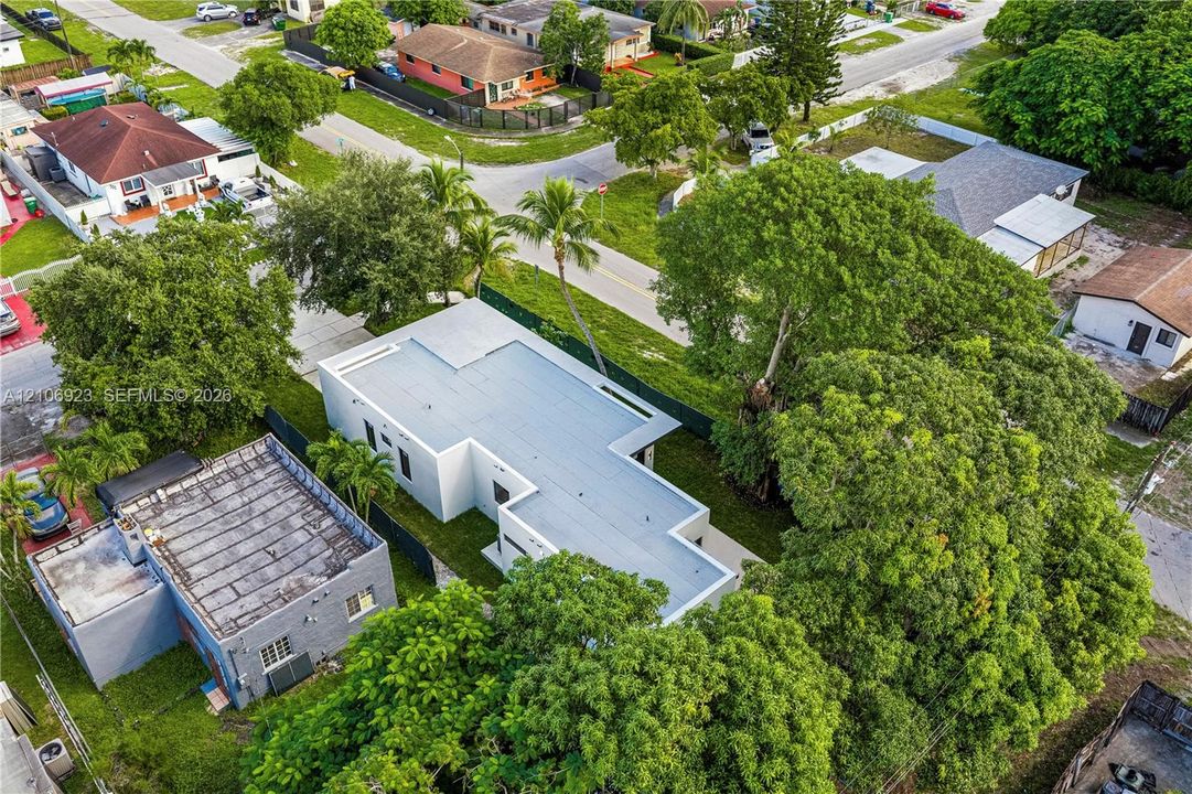 Aerial view of a rectangular property featuring a flat roof structure, concrete driveway, mature trees, and perimeter fencing