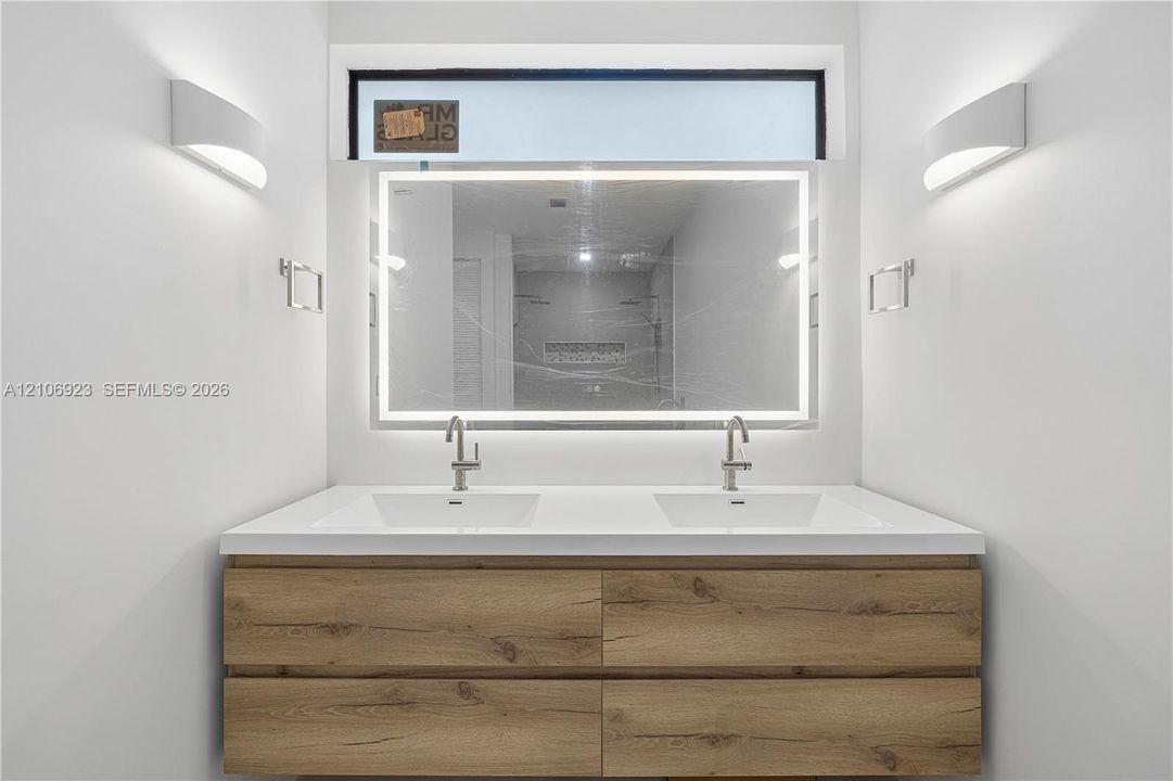 Double vanity with wood-finish cabinetry, solid surface countertop, integrated dual sinks, LED-backlit mirror, and wall-mounted sconces