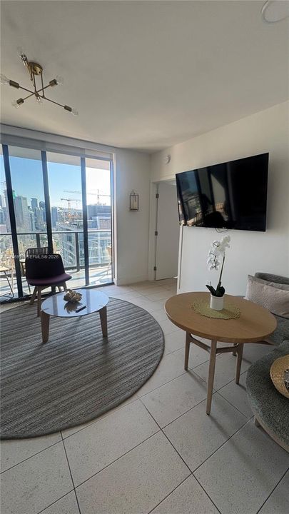 Floor-to-ceiling glass sliding doors, neutral tile flooring, multi-arm metallic chandelier, and wall-mounted television