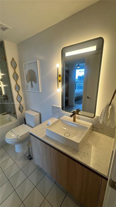 Floating vanity with a light wood-finish cabinet, stone-composite countertop, rectangular vessel sink, wall-mounted backlit mirror, and metallic finish plumbing fixtures