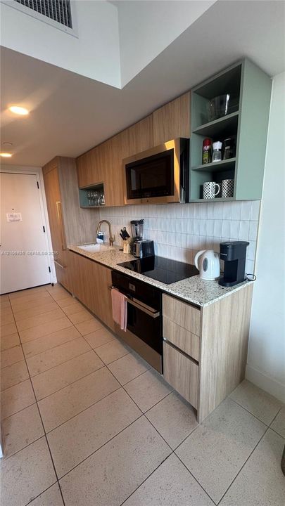 Galley kitchen featuring light wood-finish cabinetry with contrasting open shelving, stone-composite countertops, white rectangular tile backsplash, and stainless steel integrated appliances