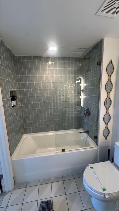 Full-height blue tile tub surround, frameless glass shower panel, recessed wall niche, brushed bronze plumbing fixtures, and geometric floor tile