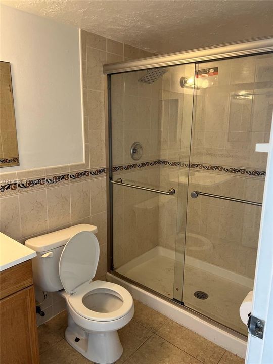 Walk-in shower featuring a glass enclosure with a sliding metal door, neutral stone-finish wall tile, an integrated shower niche, decorative tile inlay, and a built-in rainfall showerhead