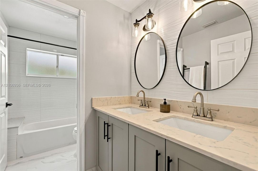Dual-sink vanity with light-toned stone countertop, shaker-style cabinetry, matte black hardware, oval mirrors, and white textured tile walls