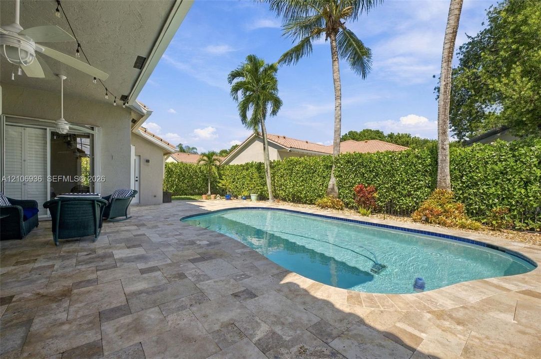 Covered patio with stone-paver flooring, built-in ceiling fan, swimming pool, and privacy-hedged perimeter